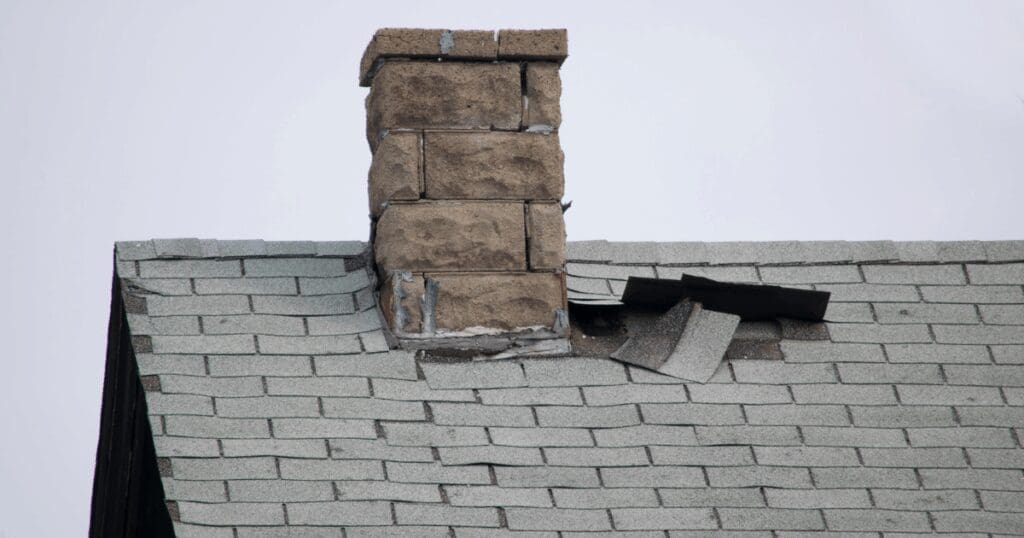 Severe winter roof damage showing torn, missing, and loose asphalt shingles around a stone chimney, leaving the roof decking exposed to leaks and requiring immediate repair.