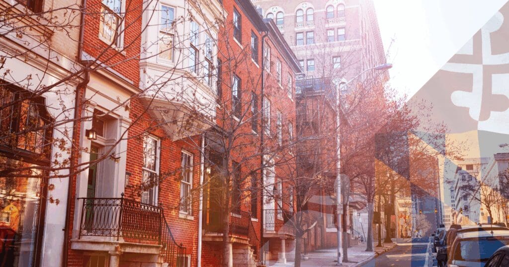 A sunny street view of historic red brick rowhouses in downtown Baltimore featuring a Maryland flag overlay. Properties in these historic districts require a specialized spring storm roof audit Baltimore specialists provide to inspect complex parapet walls and flat roof systems before the wet spring season.