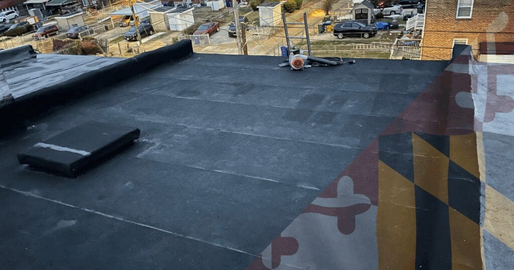 A perspective view across a large, clean dark flat residential roof membrane (Modified Bitumen or B.U.R.). An orange industrial air mover or dryer device sits near the center with wooden access stairs. Penetration flashings and more HVAC equipment are visible. A large Maryland flag watermark is overlaid, representing dedicated residential roof maintenance Baltimore properties require.